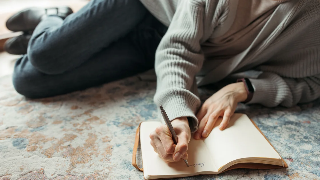 Hands writing in a notebook on a patterned rug, taking unhurried time.