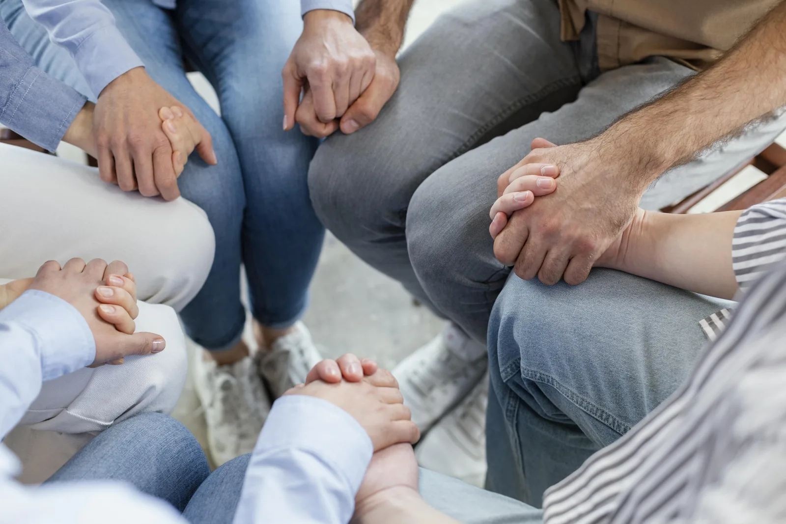 A high-angle photograph looking down into a circle of six people sitting together on the floor, their hands clasped in one another's, wearing jeans and shirts in blues, greys and white.