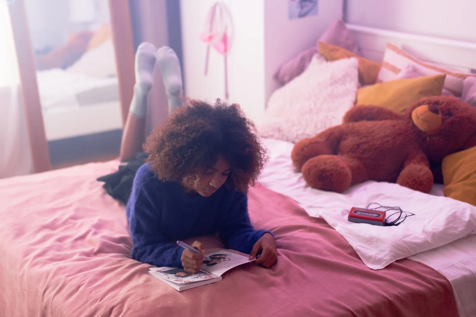 A teenager lying on her stomach on a bed, writing in a notebook, a teddy bear resting on the pillows beside her.