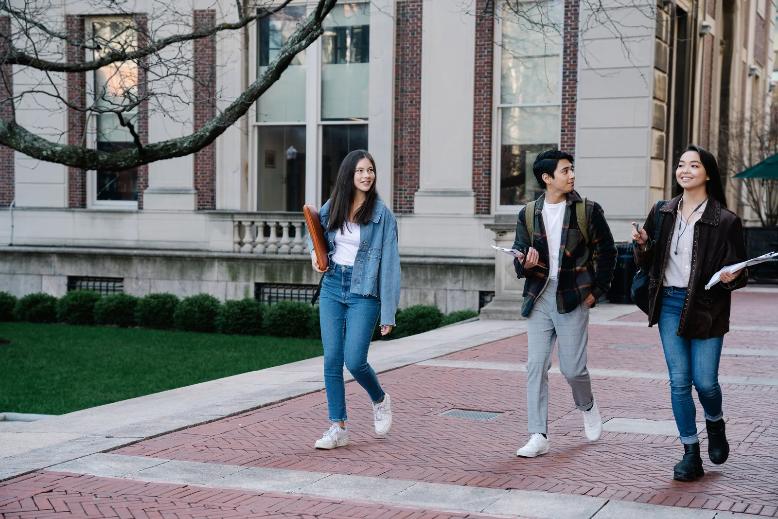 Three college students walking together along a brick campus walkway, talking, with a red-brick university building behind them.
