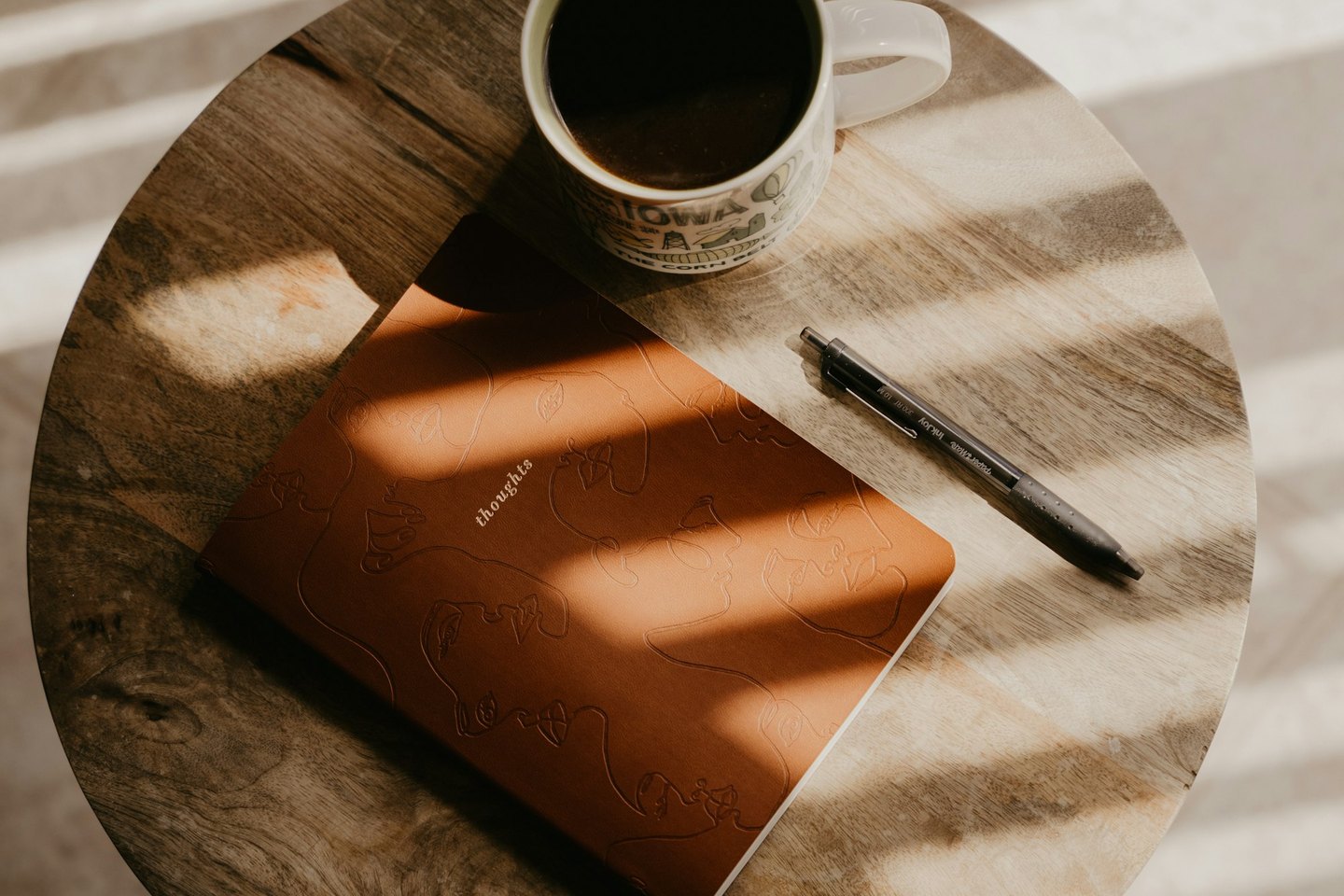 An open terracotta notebook with the word “thoughts” on its cover, resting on a round wooden table beside a mug of coffee and a pen, striped with warm light from window blinds.